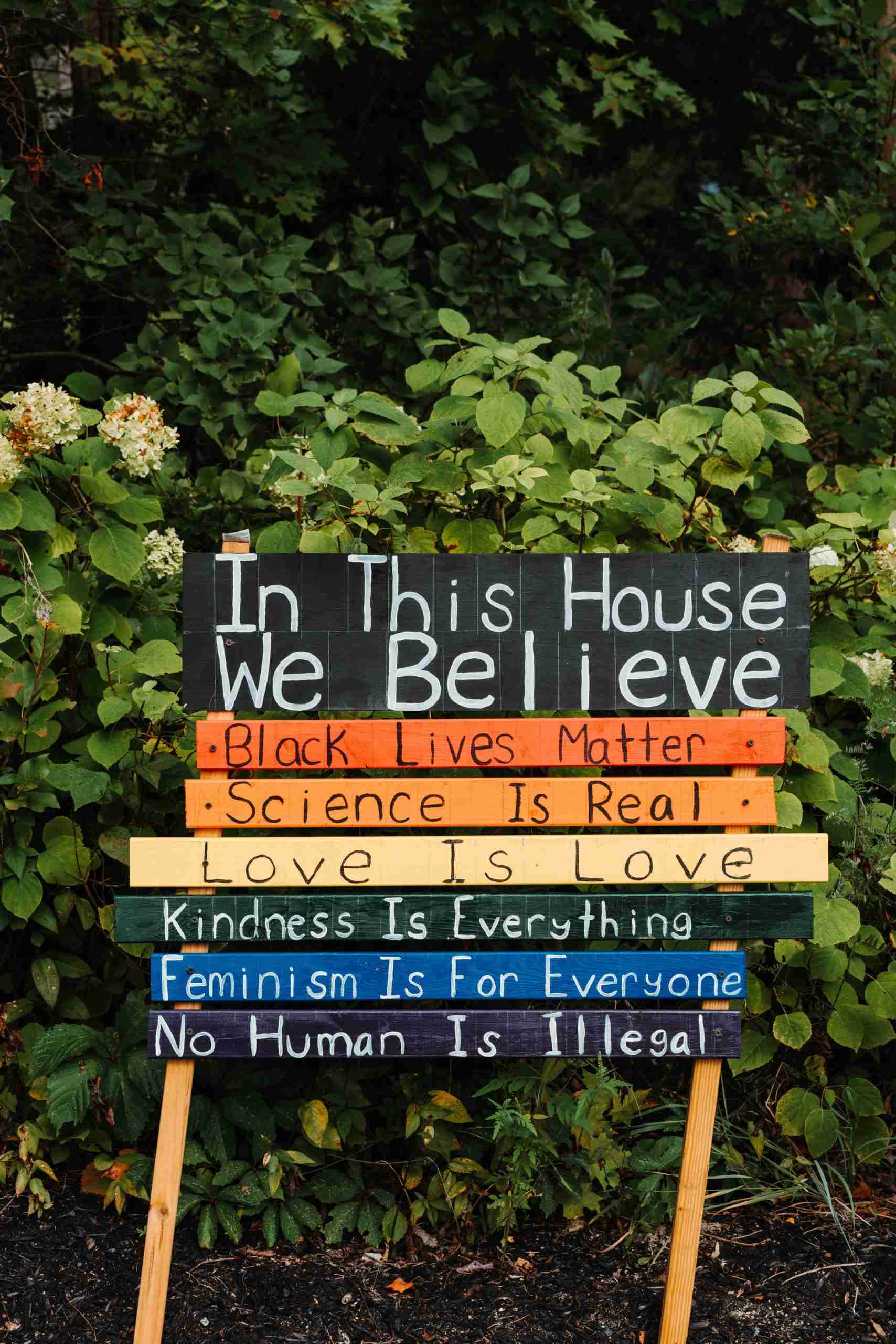 A wooden sign with rainbow colors that says, "In this house we believe, black lives matter, science is real, love is love, kindness is everything, feminism is for everyone, no human is illegal"