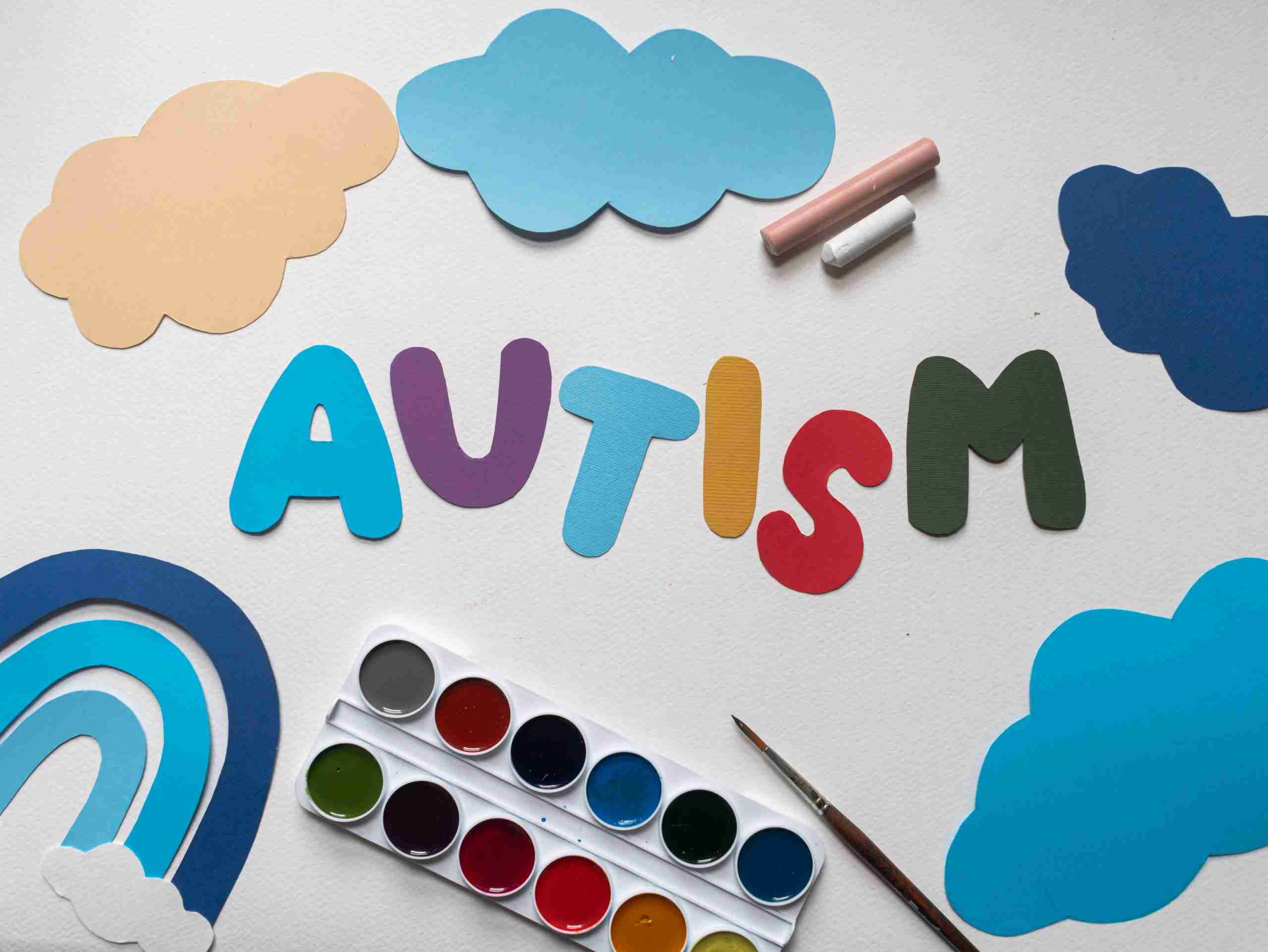 the word autism cut out of paper surrounded by paper clouds and a paper rainbow with a set of watercolors at the bottom and chalk near the top